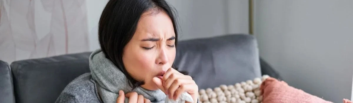 Mujer joven sentada en un sofá gris, envuelta en una bufanda y una cobija azul, tosiendo en un pañuelo desechable con expresión de malestar y los ojos cerrados.