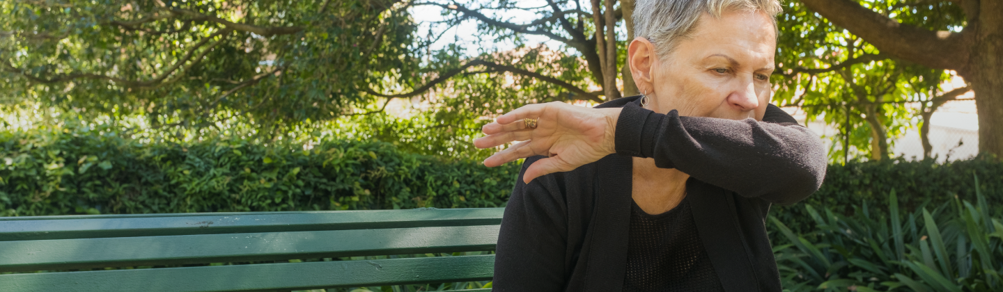 Persona adulta en la banca de un parque, tosiendo.