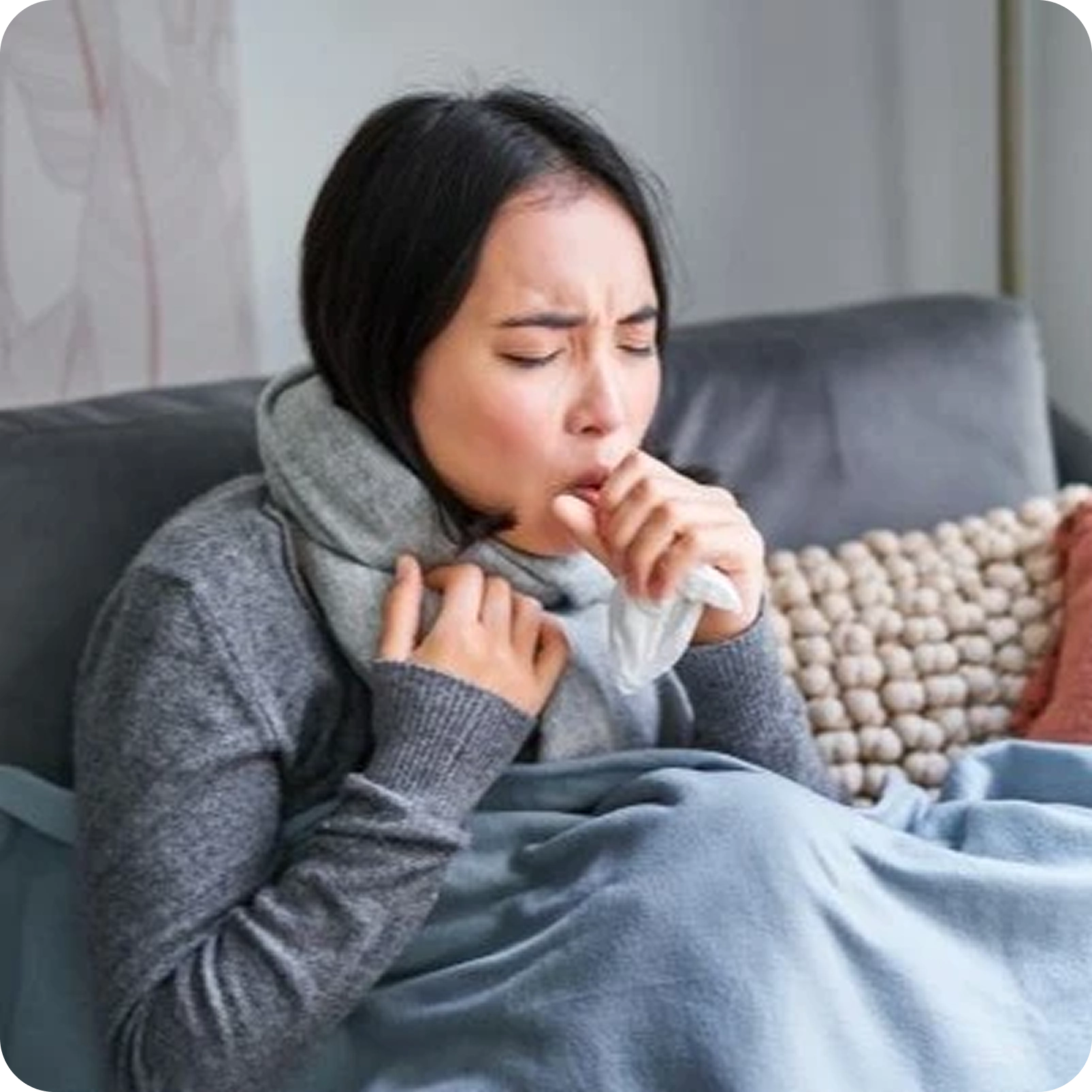 Mujer joven sentada en un sofá gris, envuelta en una bufanda y una cobija azul, tosiendo en un pañuelo desechable con expresión de malestar y los ojos cerrados.
